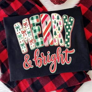May include: A black t-shirt featuring the text MERRY & BRIGHT. The word MERRY is in block letters with patterns including red bows, green plaid, red and white candy cane stripes, pink and green Christmas trees, and green polka dots.