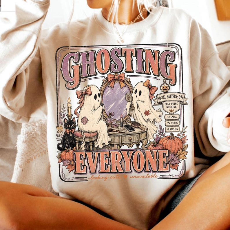 May include: A beige crewneck sweatshirt featuring a graphic print with the text GHOSTING EVERYONE. The design shows two ghosts with bows at a vanity mirror, a black cat, pumpkins, and a banner reading SOCIAL BATTERY: 0% BUSY DOING NOTHING.