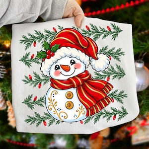May include: A white t-shirt featuring a graphic of a smiling snowman wearing a red hat with holly and a red striped scarf. The snowman has two gold buttons and gold filigree accents. The background pattern consists of green pine branches.