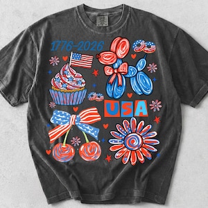 May include: A charcoal grey short-sleeve t-shirt featuring a graphic print with the text 1776-2026 and USA. The design includes a cupcake with a flag, a balloon dog, cherries with a patriotic bow, a flower, and small hearts in red, white, and blue.