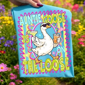 K&ouml;nnte beinhalten: Hellblaues T-Shirt mit einer Comic-Gans, die eine Sonnenbrille tr&auml;gt und eine Margarita und ein Getr&auml;nk h&auml;lt. Der Text lautet "Auntie Goose is on the Loose" in einer farbenfrohen, verspielten Schriftart.