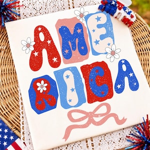 May include: A white t-shirt featuring the word AMERICA in large, colorful, blocky letters with a distressed texture. The design includes red, blue, and light pink accents, white daisy illustrations, and a pink ribbon graphic at the bottom.