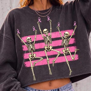 May include: A charcoal gray cropped sweatshirt featuring a graphic print of three dancing skeletons. Each skeleton is holding a small pink ribbon above its head. The design is set against three horizontal pink brushstroke stripes across the chest.