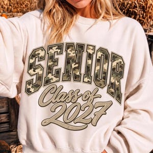 May include: A cream-coloured crewneck sweatshirt featuring the word SENIOR in a green, brown, and tan camouflage print with a black outline. Below, the text Class of 2027 is printed in a tan script font with a dark brown outline and shadow effect.