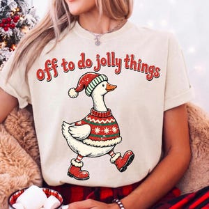 May include: A cream-colored t-shirt featuring a graphic of a white goose wearing a red and green patterned sweater, a matching Santa hat, and red boots. The text above the goose reads "off to do jolly things" in a red, distressed font.