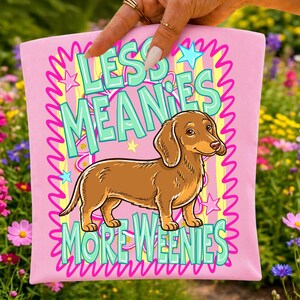 May include: A light pink fabric item featuring a brown dachshund illustration in the center. The text "LESS MEANIES MORE WEENIES" is printed in large teal letters. The design includes a yellow striped background, pink squiggly borders, and stars.