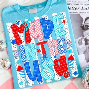 May include: A light blue t-shirt featuring the text MADE IN THE USA in large, colourful, patterned letters. The design includes red, white, and blue polka dots, checkered patterns, small blue bows, fireworks, and rocket-shaped firework graphics.