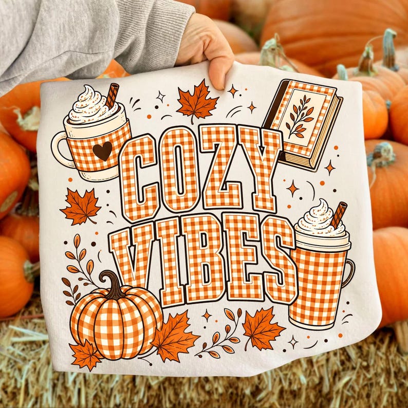 Puede incluir: Una camiseta blanca con un estampado gr&aacute;fico que incluye el texto COZY VIBES en cuadros vichy naranjas y blancos. El dise&ntilde;o presenta ilustraciones de una calabaza, dos tazas con nata montada, un libro y hojas de oto&ntilde;o en tonos naranja y marr&oacute;n.