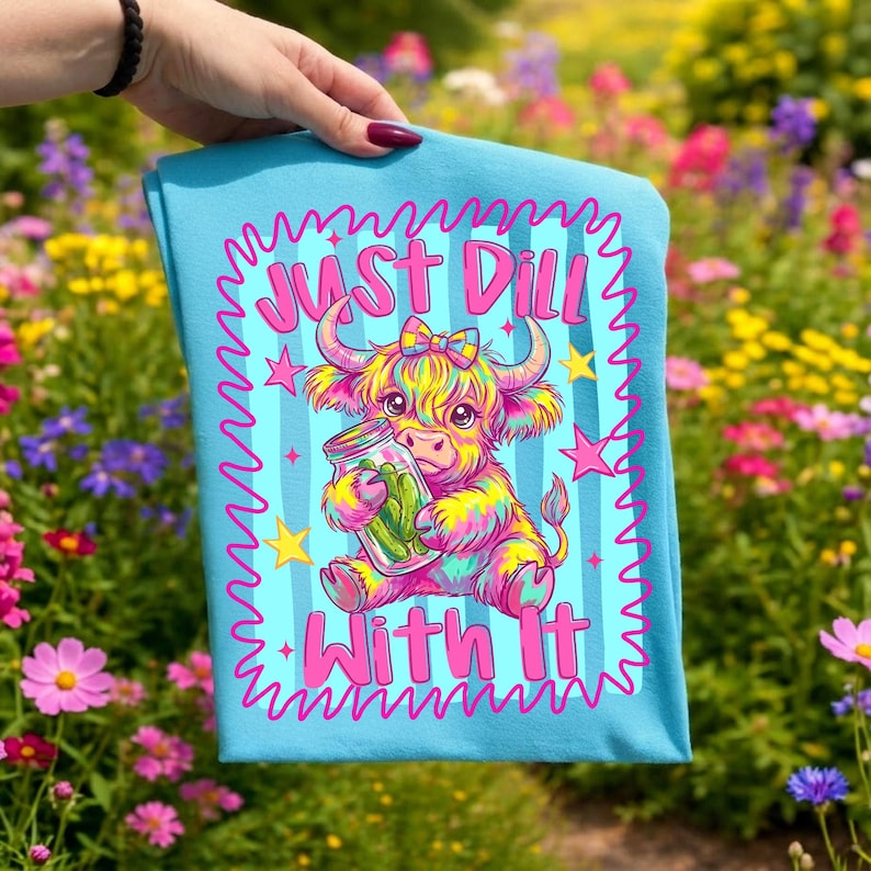 Puede incluir: Una camiseta azul claro con un gr&aacute;fico colorido de una vaca Highland peluda sosteniendo un frasco de vidrio con pepinillos. El texto "JUST DILL WITH IT" est&aacute; impreso en letras rosa brillante encima y debajo de la vaca, rodeado de estrellas rosas.