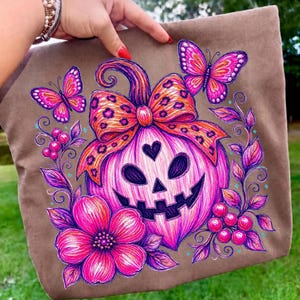 K&ouml;nnte beinhalten: Eine braune Tragetasche mit einer lebendigen Illustration eines rosa K&uuml;rbisgesichts mit herzf&ouml;rmiger Nase, gekr&ouml;nt von einer orangefarbenen Schleife mit Leopardenmuster. Das Design zeigt pinke Schmetterlinge, gro&szlig;e Blumen und kleine Beeren.