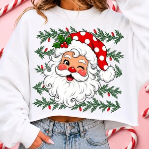Puede incluir: Una sudadera corta blanca con un gr&aacute;fico de un Santa Claus gui&ntilde;ando un ojo con un gorro rojo de lunares blancos, rodeado de ramas de pino verdes y peque&ntilde;as luces navide&ntilde;as rojas, con hojas de acebo y bayas sobre la cabeza del personaje.