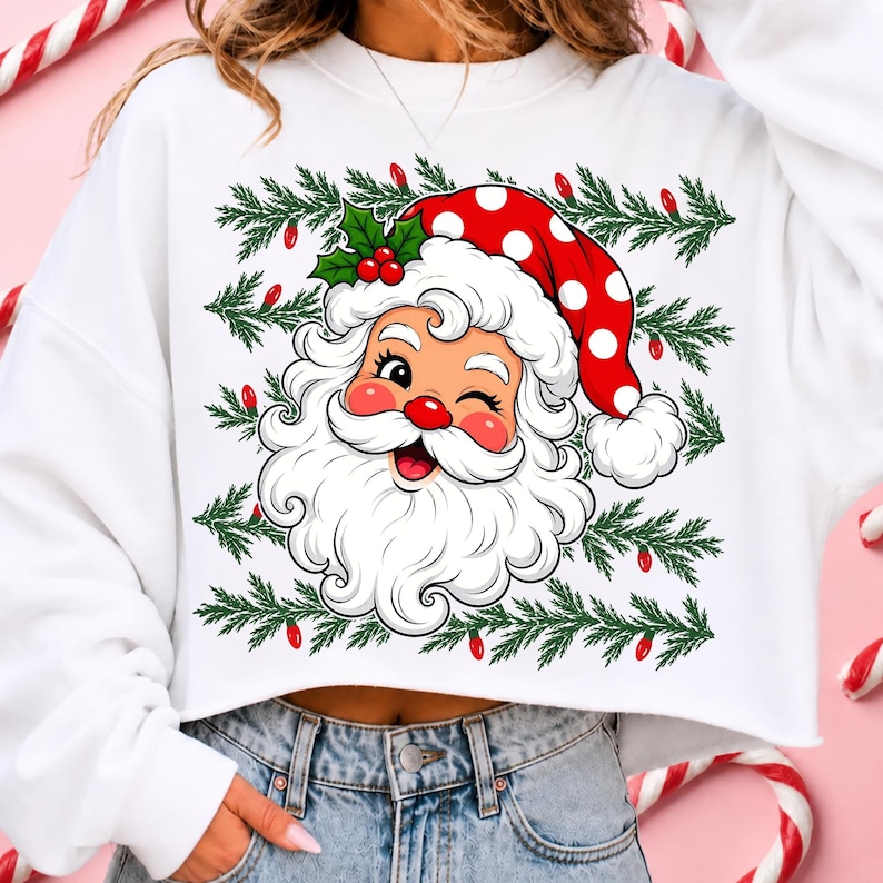 Puede incluir: Una sudadera corta blanca con un gr&aacute;fico de un Santa Claus gui&ntilde;ando un ojo con un gorro rojo de lunares blancos, rodeado de ramas de pino verdes y peque&ntilde;as luces navide&ntilde;as rojas, con hojas de acebo y bayas sobre la cabeza del personaje.