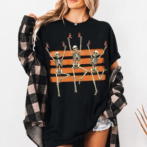 May include: A black t-shirt featuring a graphic of three skeletons dancing with their arms raised. Each skeleton holds a small pink ribbon. Behind the skeletons are three horizontal orange brushstroke stripes. The shirt is worn with a black and tan plaid shirt.