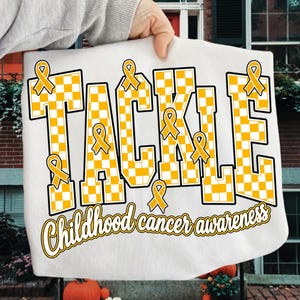 May include: A white t-shirt featuring the word TACKLE in large, yellow and white checkered block letters. Several yellow awareness ribbons are integrated into the design. Below, the text Childhood cancer awareness is written in yellow script.