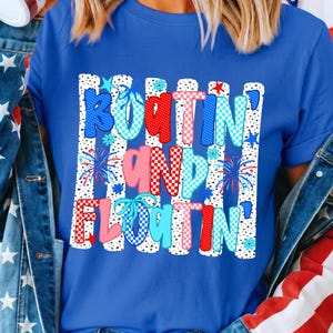 Op de afbeelding: Een koningsblauw T-shirt met korte mouwen en een grafische print met de tekst "BOATIN' AND FLOATIN'" in rood, wit en lichtblauw geblokte en gestippelde patronen, geaccentueerd met kleine blauwe vuurwerk-, ster- en bloemillustraties.