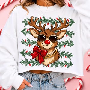 Puede incluir: Una sudadera blanca de manga larga con un gr&aacute;fico de un reno marr&oacute;n que lleva gafas de sol oscuras, un lazo rojo grande y un collar de perlas. El fondo del gr&aacute;fico incluye filas horizontales de ramas de pino verdes y peque&ntilde;as bombillas rojas.