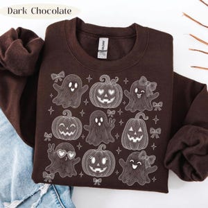 Puede incluir: Una sudadera de cuello redondo en color marr&oacute;n chocolate oscuro con un gr&aacute;fico de l&iacute;neas blancas que muestra varios fantasmas y calabazas decorados con lazos y estrellas. El texto "Dark Chocolate" aparece en la esquina superior izquierda.