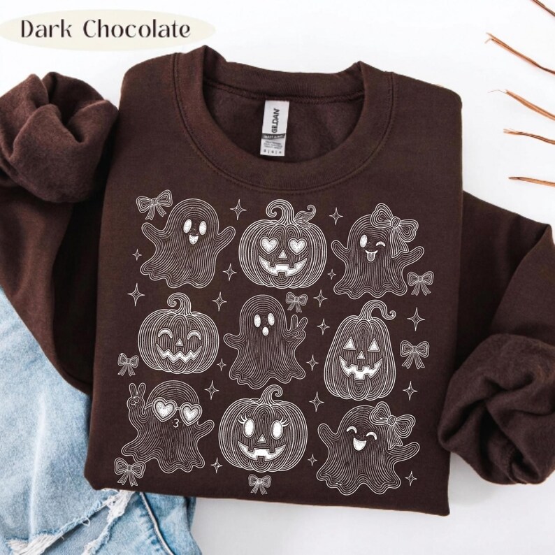 Puede incluir: Una sudadera de cuello redondo en color marr&oacute;n chocolate oscuro con un gr&aacute;fico de l&iacute;neas blancas que muestra varios fantasmas y calabazas decorados con lazos y estrellas. El texto "Dark Chocolate" aparece en la esquina superior izquierda.