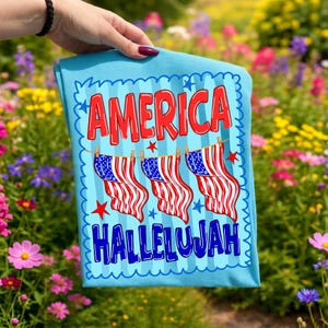 Puede incluir: Una camiseta azul claro con el texto rojo AMERICA en la parte superior y el texto azul HALLELUJAH en la parte inferior. Entre las palabras hay tres banderas estadounidenses colgadas en un tendedero con peque&ntilde;as estrellas rojas dispersas.