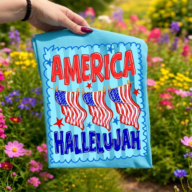 Puede incluir: Una camiseta azul claro con el texto rojo AMERICA en la parte superior y el texto azul HALLELUJAH en la parte inferior. Entre las palabras hay tres banderas estadounidenses colgadas en un tendedero con peque&ntilde;as estrellas rojas dispersas.