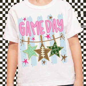 May include: A white t-shirt featuring a graphic print with the text "GAME DAY" in pink letters. The design includes light blue vertical stripes, a brown twine string with wooden clothespins, and three stars displaying football-themed patterns.