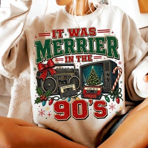 Może przedstawiać: Kremowa bluza z grafiką i napisem IT WAS MERRIER IN THE 90'S. Wz&oacute;r przedstawia zabytkowy radiomagnetofon z czerwoną kokardą, telewizor z choinką, kasety magnetofonowe oraz kolorowe lampki świąteczne rozmieszczone wok&oacute;ł element&oacute;w graficznych.