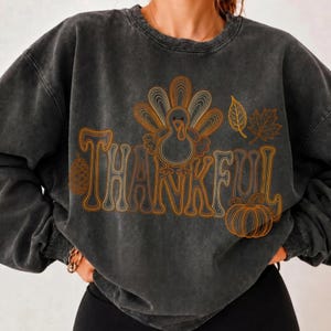 May include: A charcoal gray mineral-washed crewneck sweatshirt featuring a graphic print in orange and gold line art. The design includes the word THANKFUL, a turkey, a pinecone, a pumpkin, and autumn leaves arranged across the chest area.