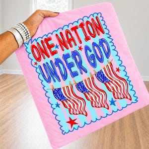 May include: A light pink square fabric item featuring the text ONE NATION UNDER GOD in red and blue letters. Below the text, three American flags hang from a clothesline. The design is enclosed in a blue scalloped border with small red stars.