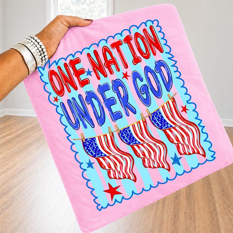 May include: A light pink square fabric item featuring the text ONE NATION UNDER GOD in red and blue letters. Below the text, three American flags hang from a clothesline. The design is enclosed in a blue scalloped border with small red stars.