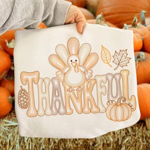 May include: A white sweatshirt featuring a brown line art graphic of a turkey, a pinecone, two leaves, and a pumpkin. The word THANKFUL is written in large, stylized, brown-outlined letters. The design is centered on the front of the garment.