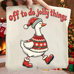 May include: A cream-colored t-shirt featuring a graphic of a white goose wearing a red and green patterned sweater, a matching Santa hat, and red boots. The text "off to do jolly things" is printed in red above the goose in a playful font.