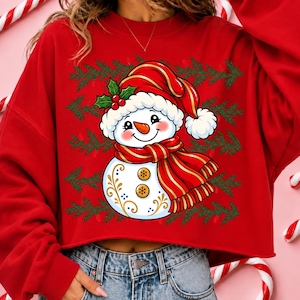 May include: A red long-sleeve sweatshirt featuring a graphic of a smiling snowman wearing a red hat with holly and a red striped scarf. The snowman has two gold buttons and gold filigree details on its body, set against a background of green pine branches.