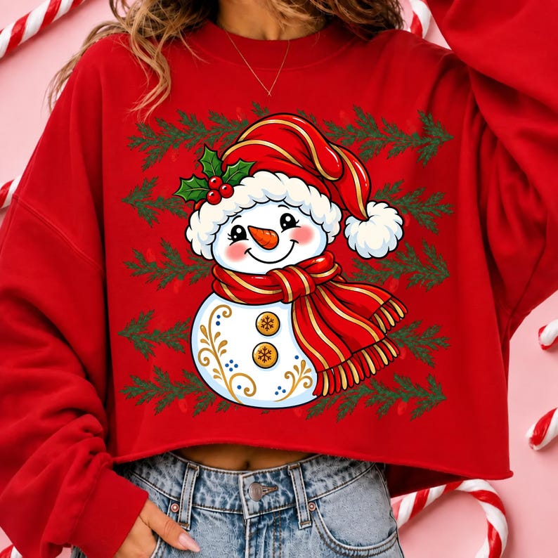 May include: A red long-sleeve sweatshirt featuring a graphic of a smiling snowman wearing a red hat with holly and a red striped scarf. The snowman has two gold buttons and gold filigree details on its body, set against a background of green pine branches.