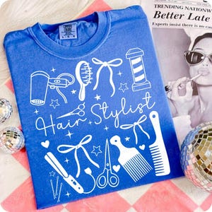 May include: A blue t-shirt with a white graphic design featuring hairstyling tools and the words "Hair Stylist." The design includes a hair dryer, scissors, combs, and a barber pole. The shirt is a crew neck.
