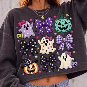 K&ouml;nnte beinhalten: Ein anthrazitfarbenes bauchfreies Sweatshirt mit einem Grafikdruck aus drei Geistern, zwei K&uuml;rbissen und vier Schleifen. Die Geister sind wei&szlig; und hellviolett, tragen rosa Schleifen und halten kleine K&uuml;rbisse. Das Design enth&auml;lt bunte Sterne.