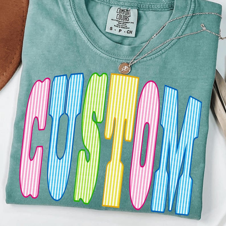 May include: A sage green short-sleeve t-shirt with a Comfort Colors label at the neck. The front features the word CUSTOM in large, bold, vertical-striped letters in alternating colors of pink, blue, green, and yellow. A thin silver chain necklace rests on the shirt.