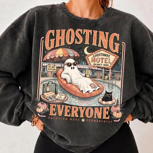 Puede incluir: Una sudadera gris carb&oacute;n con lavado mineral que presenta un estampado gr&aacute;fico con el texto GHOSTING EVERYONE. El dise&ntilde;o muestra un fantasma con gafas de sol en un flotador, un gato negro, un letrero de GHOSTAWAY MOTEL y calabazas.