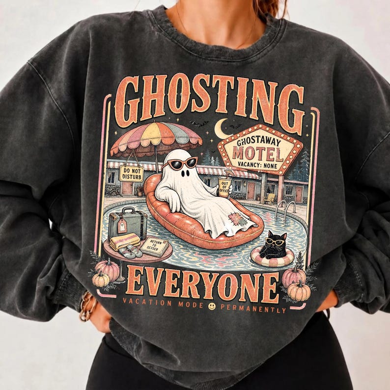 Puede incluir: Una sudadera gris carb&oacute;n con lavado mineral que presenta un estampado gr&aacute;fico con el texto GHOSTING EVERYONE. El dise&ntilde;o muestra un fantasma con gafas de sol en un flotador, un gato negro, un letrero de GHOSTAWAY MOTEL y calabazas.