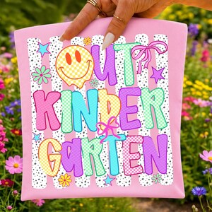Puede incluir: Un art&iacute;culo de tela rosa claro con un estampado gr&aacute;fico colorido con el texto OUT KINDERGARTEN. El dise&ntilde;o incluye una cara sonriente a cuadros amarillos, letras rosas y verdes con patrones de puntos, peque&ntilde;as estrellas, flores y lazos.