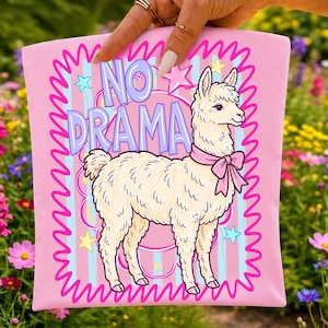 Peut inclure: Un carr&eacute; de tissu rose clair illustr&eacute; d'un lama cr&egrave;me portant un n&oelig;ud rose autour du cou. Le texte NO DRAMA est imprim&eacute; en violet au-dessus du lama. Le motif comprend des rayures verticales bleues, des &eacute;toiles roses et une bordure ondul&eacute;e.