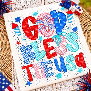 K&ouml;nnte beinhalten: Ein wei&szlig;es rechteckiges Schild mit dem Text GOD BLESS THE USA in roten, blauen und rosafarbenen Karomustern und Punkten. Das Design enth&auml;lt Feuerwerksillustrationen, kleine Sterne und Blumen sowie eine patriotische Haarspange an der Ecke.