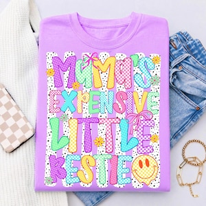 May include: A light purple t-shirt featuring the text "MAMA'S EXPENSIVE LITTLE BESTIE" in colorful, patterned block letters. The design includes pink bows, small yellow and green flowers, and a yellow smiley face, all set against vertical white stripes.