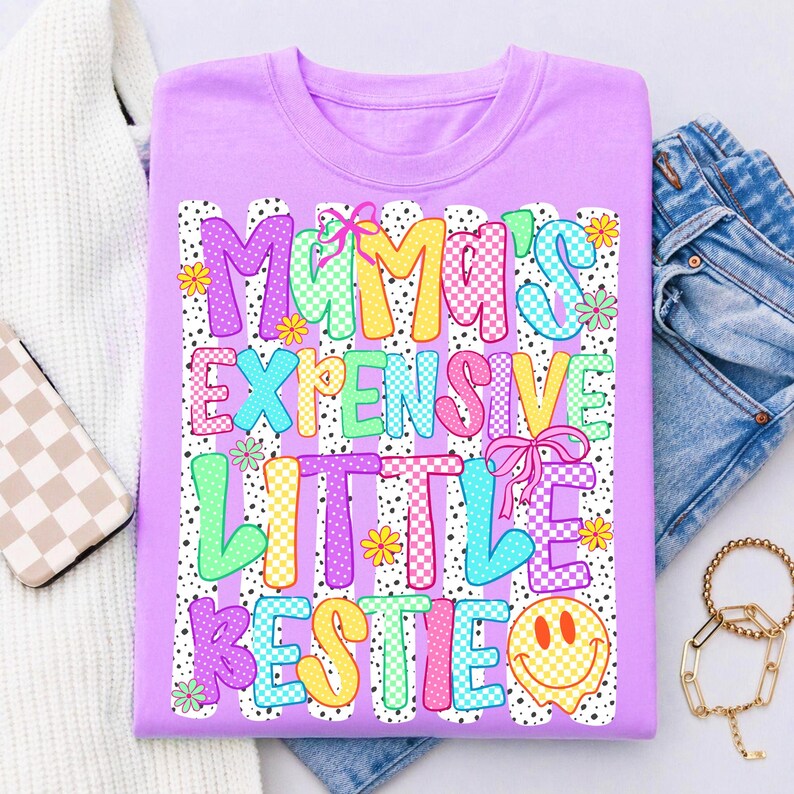 May include: A light purple t-shirt featuring the text "MAMA'S EXPENSIVE LITTLE BESTIE" in colorful, patterned block letters. The design includes pink bows, small yellow and green flowers, and a yellow smiley face, all set against vertical white stripes.