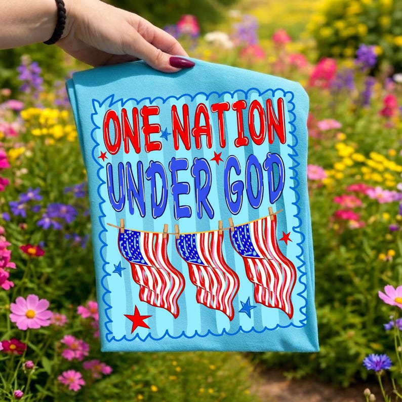 May include: A light blue t-shirt featuring the text ONE NATION UNDER GOD in red and blue letters. Below the text, three American flags hang from a clothesline. The design is framed by a scalloped border with small red and blue stars.