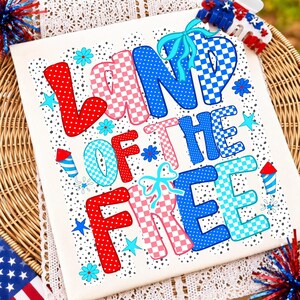 May include: A white t-shirt featuring the text LAND OF THE FREE in large, colorful, patterned letters. The design includes red, white, and blue polka dots and checkered patterns, accented with light blue bows, small flowers, stars, and firework icons.