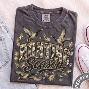 May include: A charcoal gray crew neck t-shirt featuring a graphic print of three flying ducks and oak leaves. The text "HUNTING Season" is printed in a camouflage pattern and black script. The shirt has a Comfort Colors brand neck label.