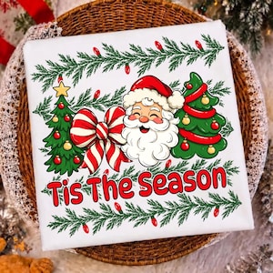 Puede incluir: Una camiseta blanca con un gr&aacute;fico de Santa Claus, dos &aacute;rboles de Navidad decorados y un gran lazo a rayas rojas y blancas. El texto 'Tis The Season' est&aacute; impreso en rojo debajo del gr&aacute;fico, enmarcado por ramas de pino verdes y luces rojas.