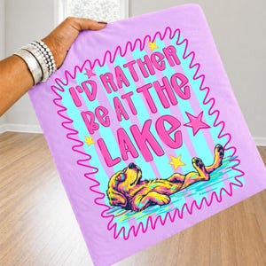 May include: A light purple fabric square featuring a graphic print with the text I'D RATHER BE AT THE LAKE in pink letters. The design includes a yellow dog wearing sunglasses floating in blue water, surrounded by a pink wavy border and yellow stars.
