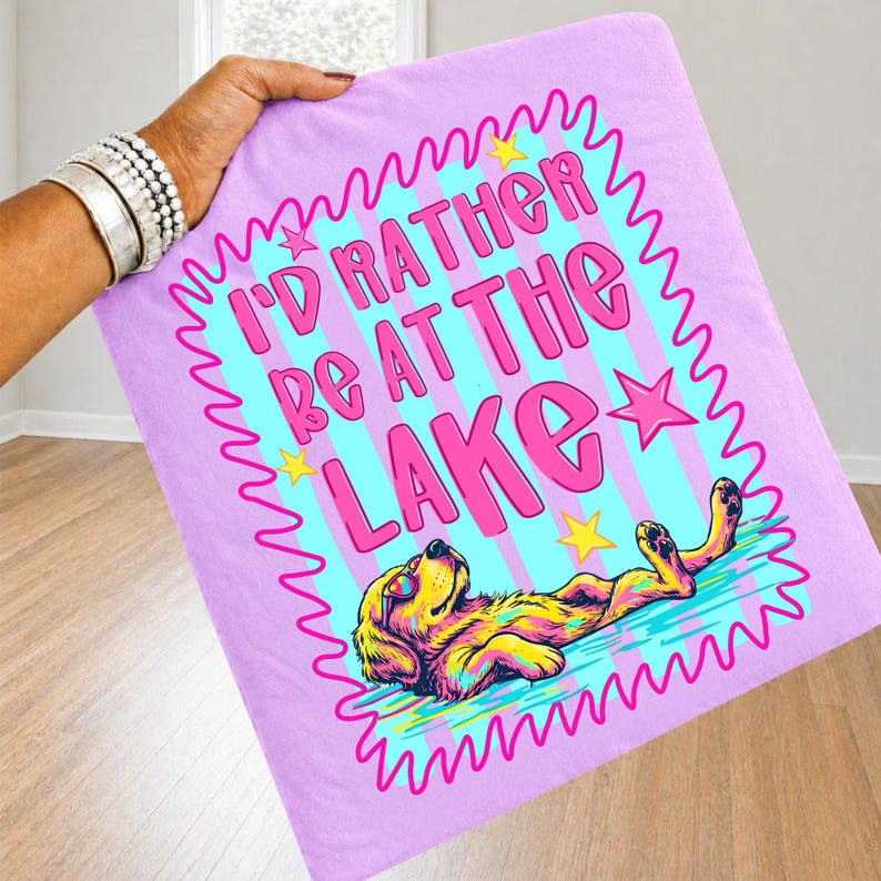 May include: A light purple fabric square featuring a graphic print with the text I'D RATHER BE AT THE LAKE in pink letters. The design includes a yellow dog wearing sunglasses floating in blue water, surrounded by a pink wavy border and yellow stars.