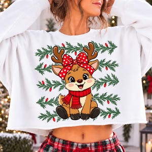 May include: A white cropped sweatshirt featuring a central graphic of a brown cartoon reindeer with a red and white polka dot bow and a red striped scarf. The background of the graphic consists of horizontal green pine branches and red lights.